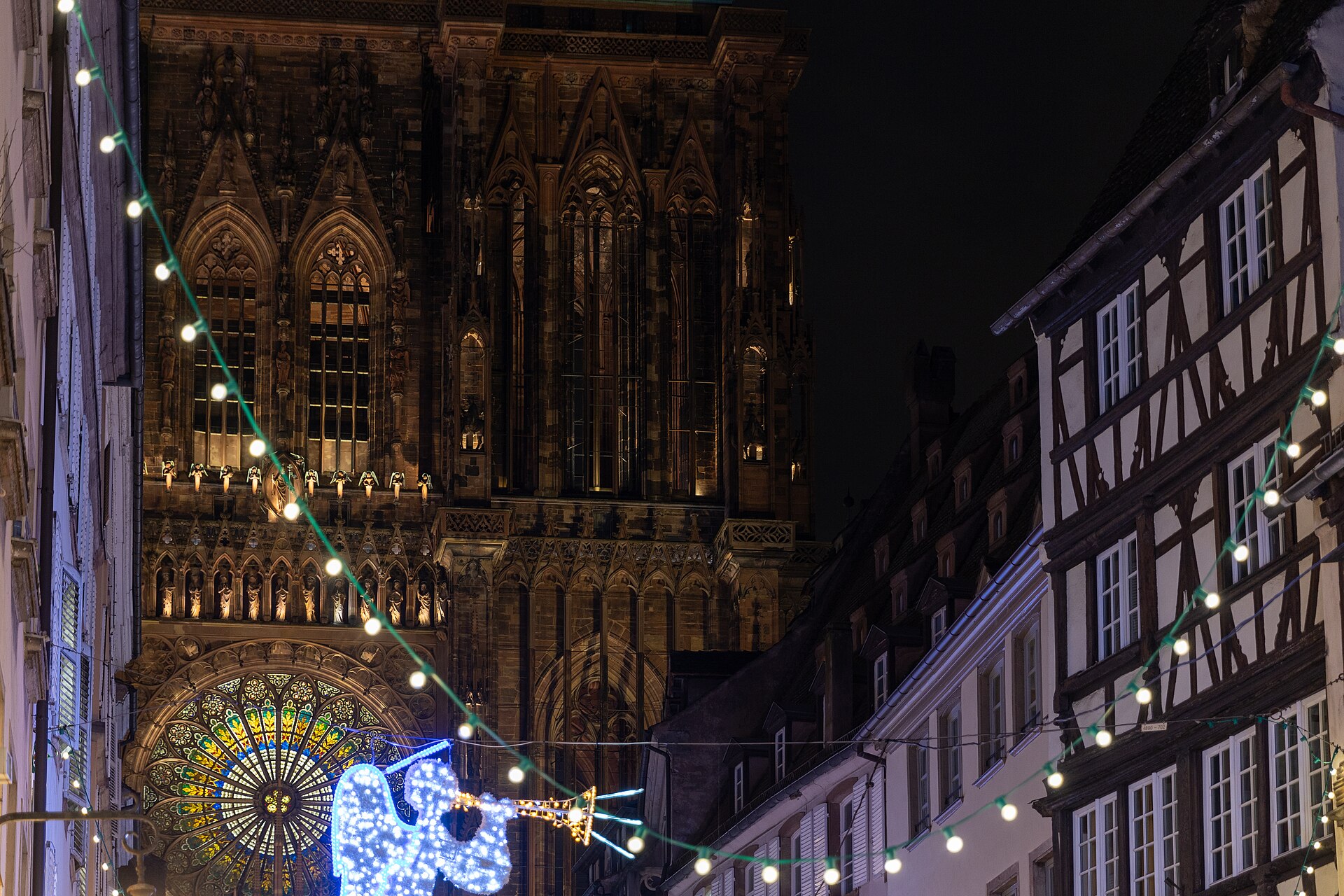 La rosace de la cathédrale de Strasbourg illuminée, un ange de lumière et des guirlandes au-dessus de la rue, la nuit, pendant le marché de Noël
